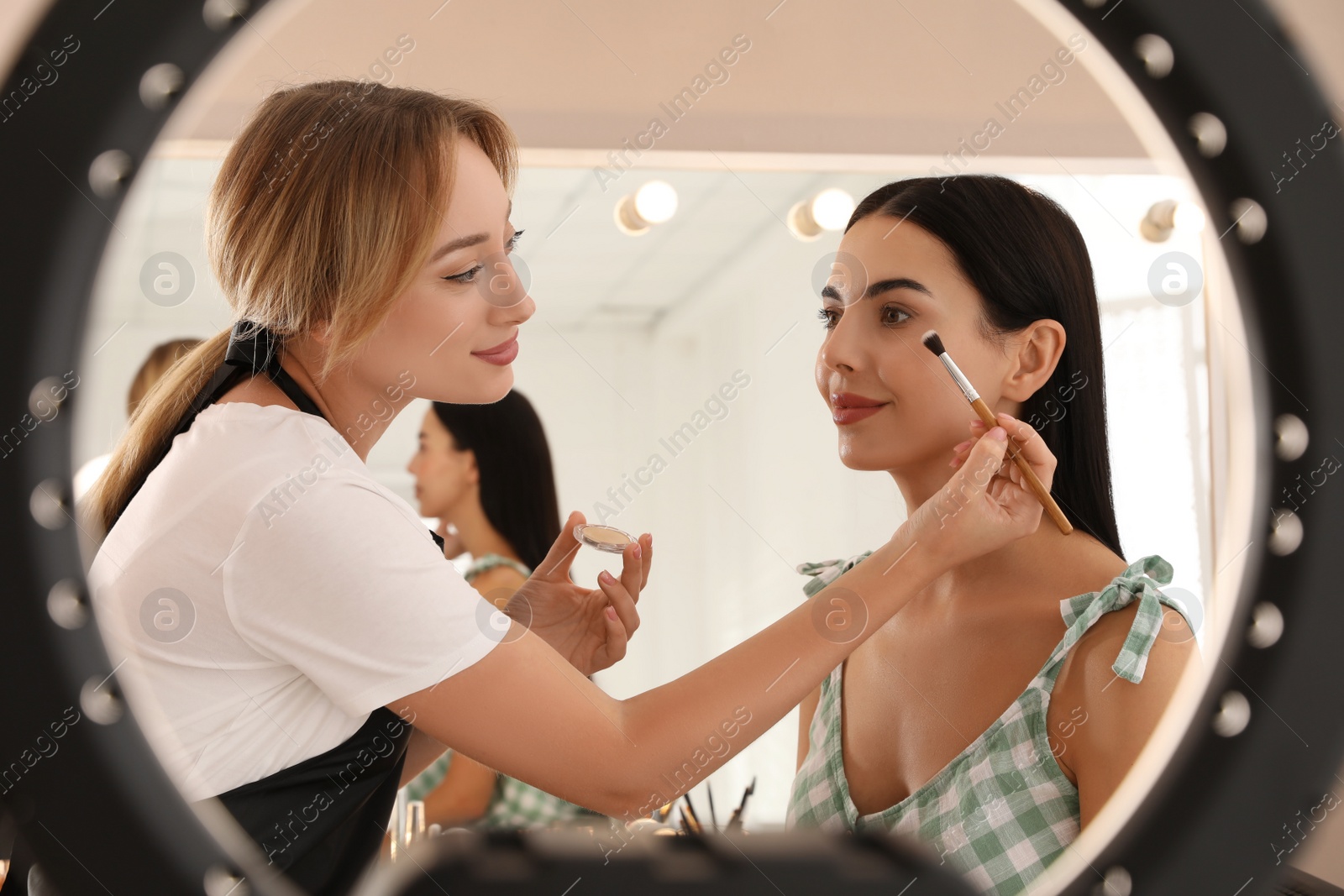 Photo of Professional makeup artist working with beautiful young woman in salon, view through ring lamp