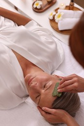 Photo of Woman receiving facial massage with jade gua sha tool in beauty salon, above view
