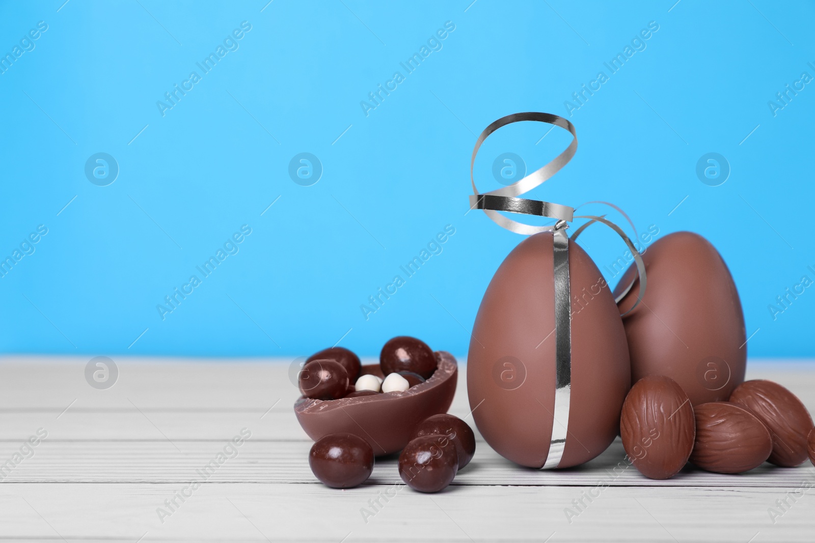 Photo of Delicious chocolate eggs and candies on white wooden table against light blue background. Space for text