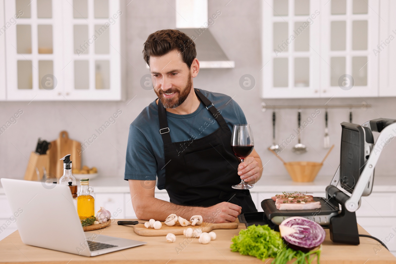 Photo of Man with glass of wine making dinner while watching online cooking course via laptop in kitchen