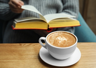 Photo of Woman reading book at table, focus on cup of coffee
