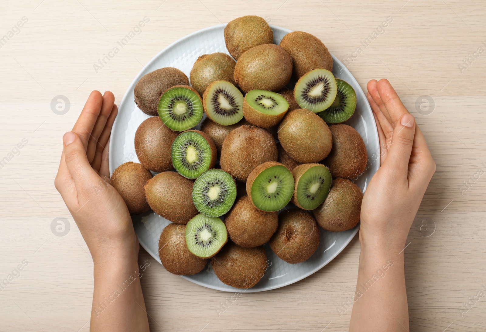 Photo of Woman holding plate with fresh ripe kiwis at light wooden table, top view