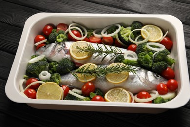 Raw fish with vegetables and lemon in baking dish on black wooden table