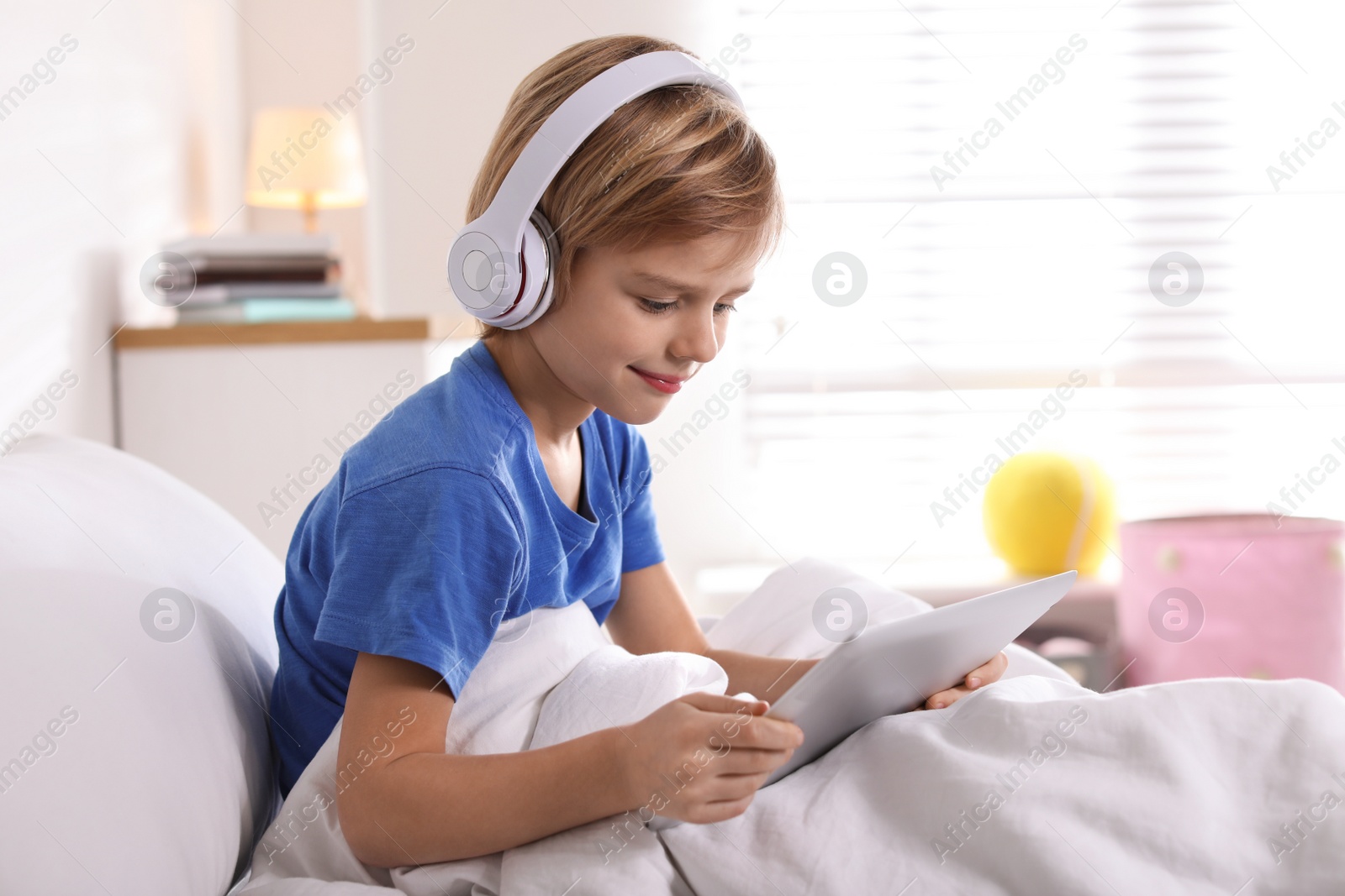Photo of Cute little boy with headphones and tablet listening to audiobook in bed at home
