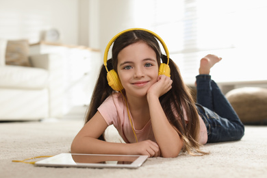 Photo of Cute little girl with headphones and tablet listening to audiobook at home