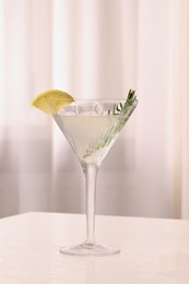 Elegant martini glass with fresh cocktail, rosemary and lemon slice on white textured table indoors