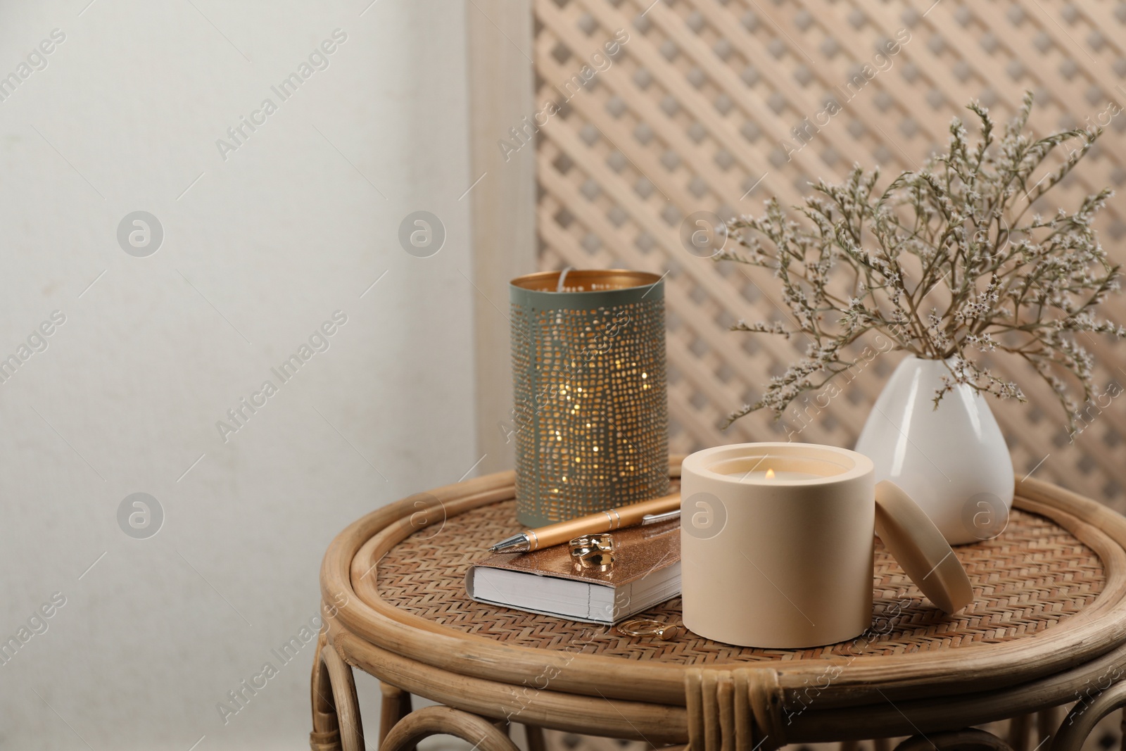 Photo of Burning soy candle, notebook and stylish accessories on wicker table indoors. Space for text