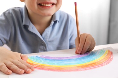 Photo of Little boy drawing rainbow with pencil at white table indoors, closeup. Child`s art