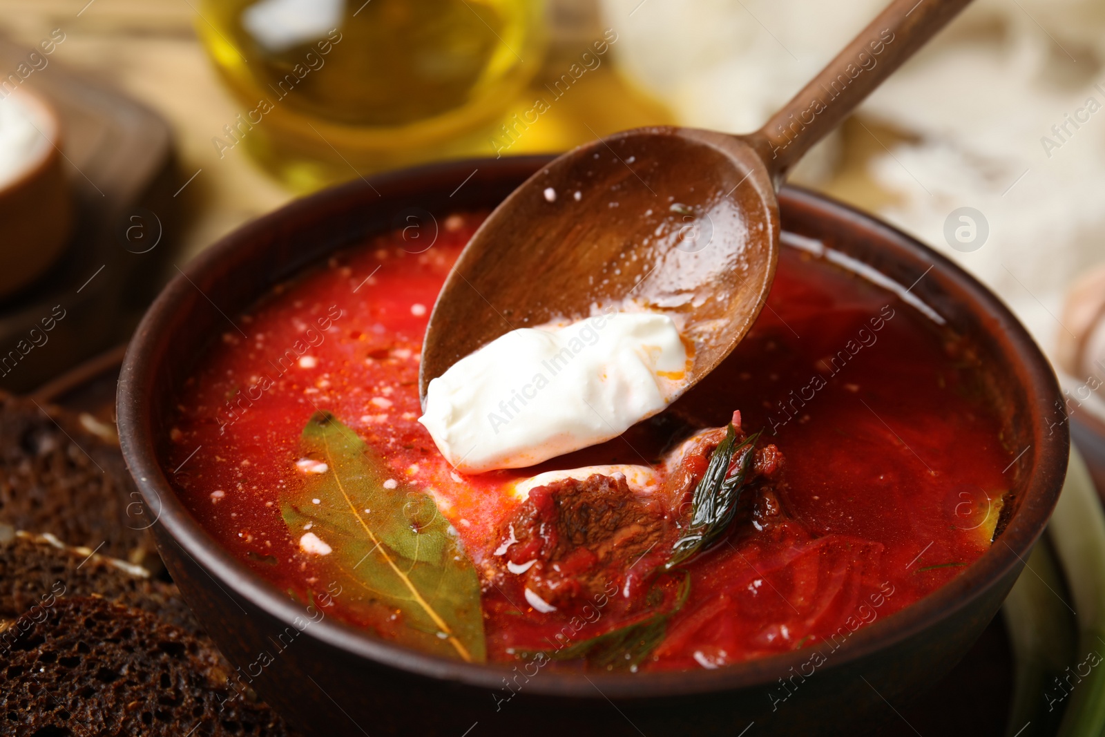 Photo of Putting sour cream into clay bowl with Ukrainian borsch on table, closeup