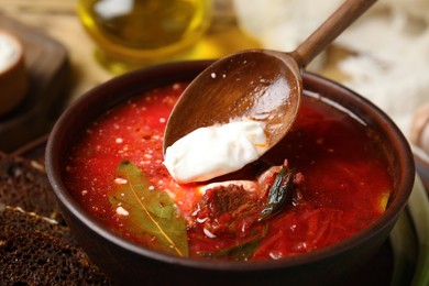 Putting sour cream into clay bowl with Ukrainian borsch on table, closeup