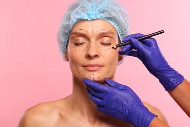 Doctor with pencil preparing patient for cosmetic surgery operation on pink background, closeup