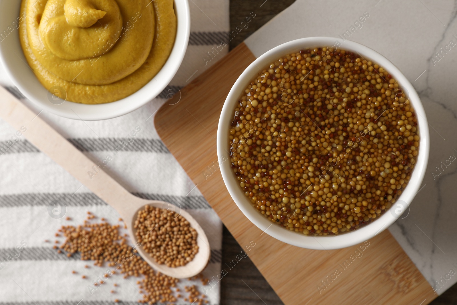 Photo of Bowls and spoon of whole grain mustard on wooden table, flat lay