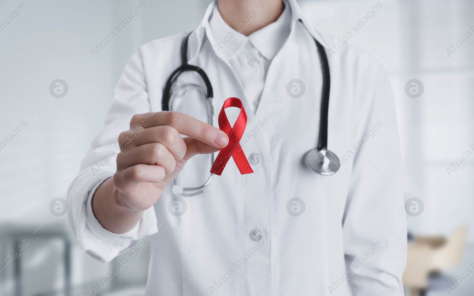 Image of Doctor holding red awareness ribbon on blurred background, closeup. World AIDS disease day