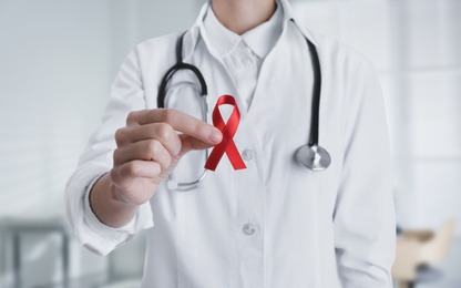 Doctor holding red awareness ribbon on blurred background, closeup. World AIDS disease day