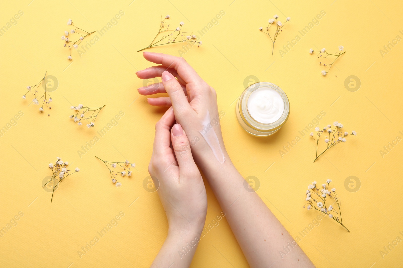 Photo of Woman applying hand cream and flowers on yellow background, top view