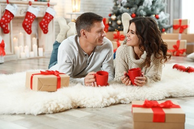 Happy couple with cups of hot drink on floor in room decorated for Christmas