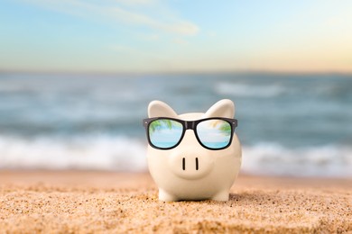 Vacation savings. Piggy bank on sandy beach near sea. Reflection of palm leaves in sunglasses