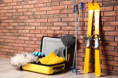 Photo of Suitcase with clothes, camera and skis on floor against brick wall, space for text. Winter vacation