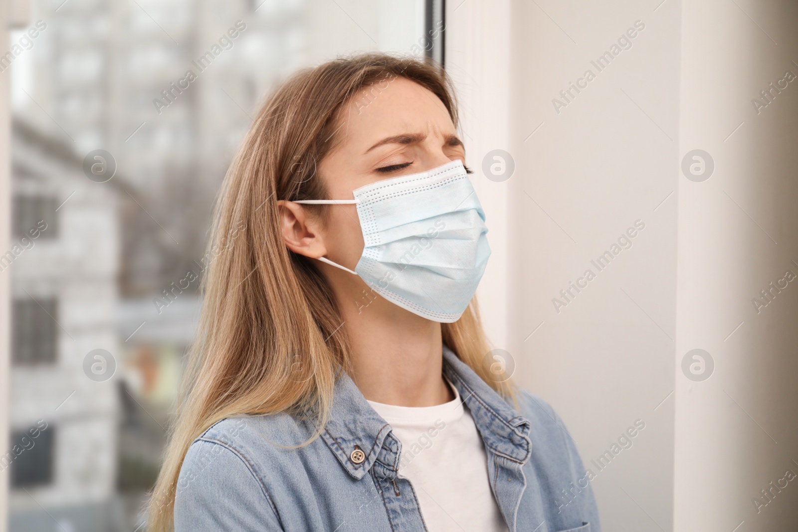 Photo of Sad woman in protective mask near window indoors. Staying at home during coronavirus pandemic