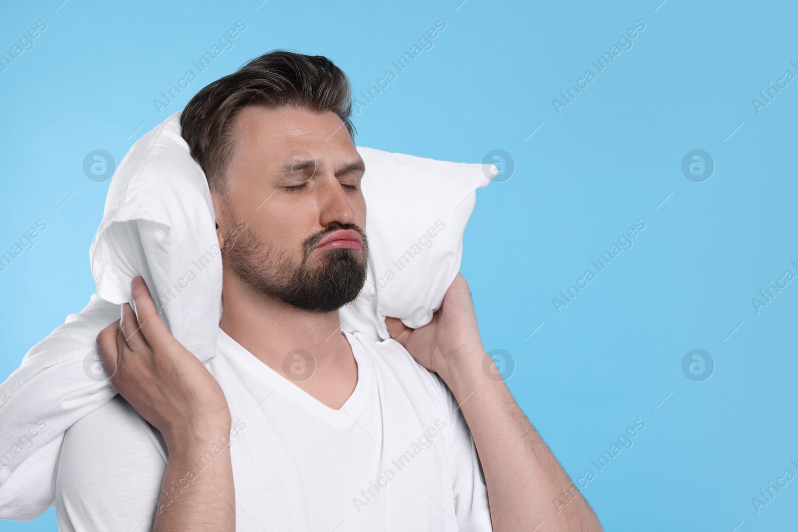 Photo of Unhappy man with pillow on light blue background, space for text. Insomnia problem