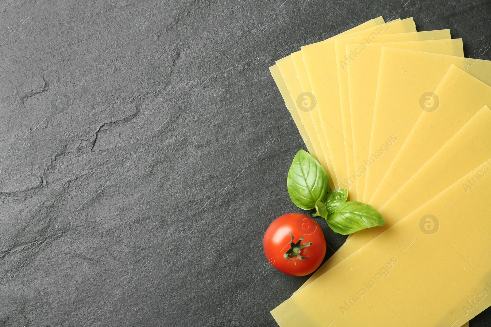 Photo of Uncooked lasagna sheets, tomato and basil on dark background, flat lay. Space for text