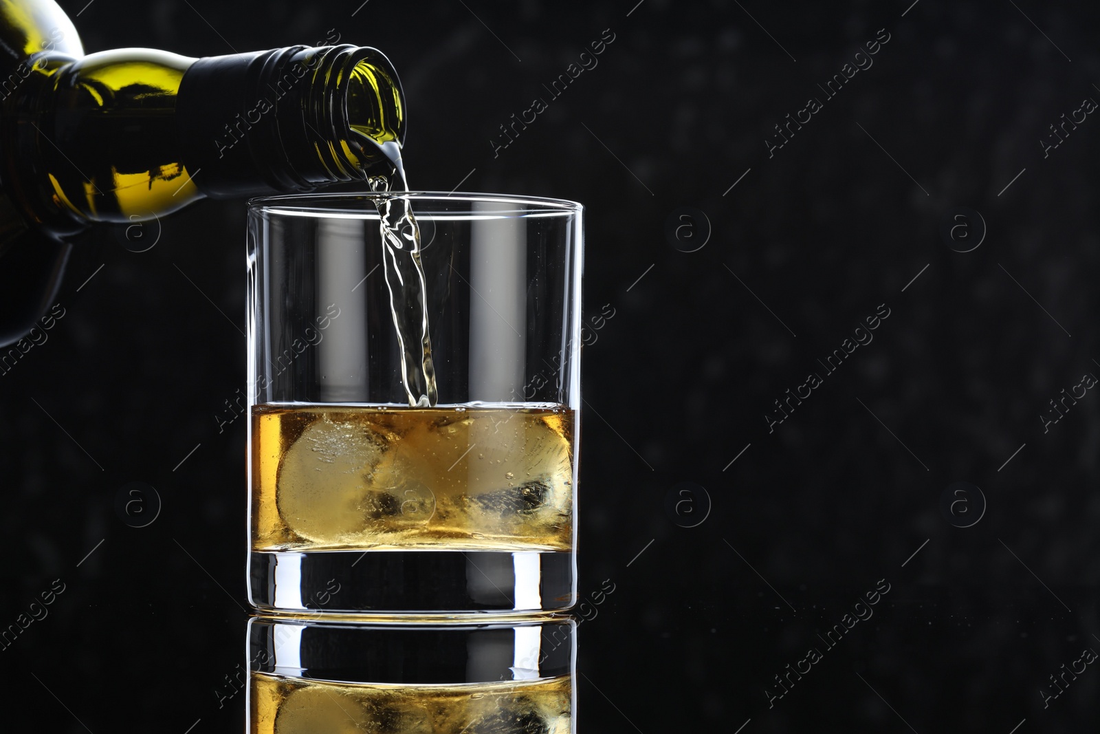 Photo of Pouring tasty whiskey from bottle into glass at mirror table against black background, closeup. Space for text