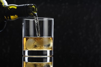 Photo of Pouring tasty whiskey from bottle into glass at mirror table against black background, closeup. Space for text