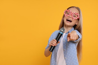 Photo of Cute little girl with funny glasses and microphone singing on yellow background, space for text