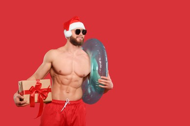 Photo of Muscular young man in Santa hat and sunglasses with inflatable ring and gift box on red background, space for text