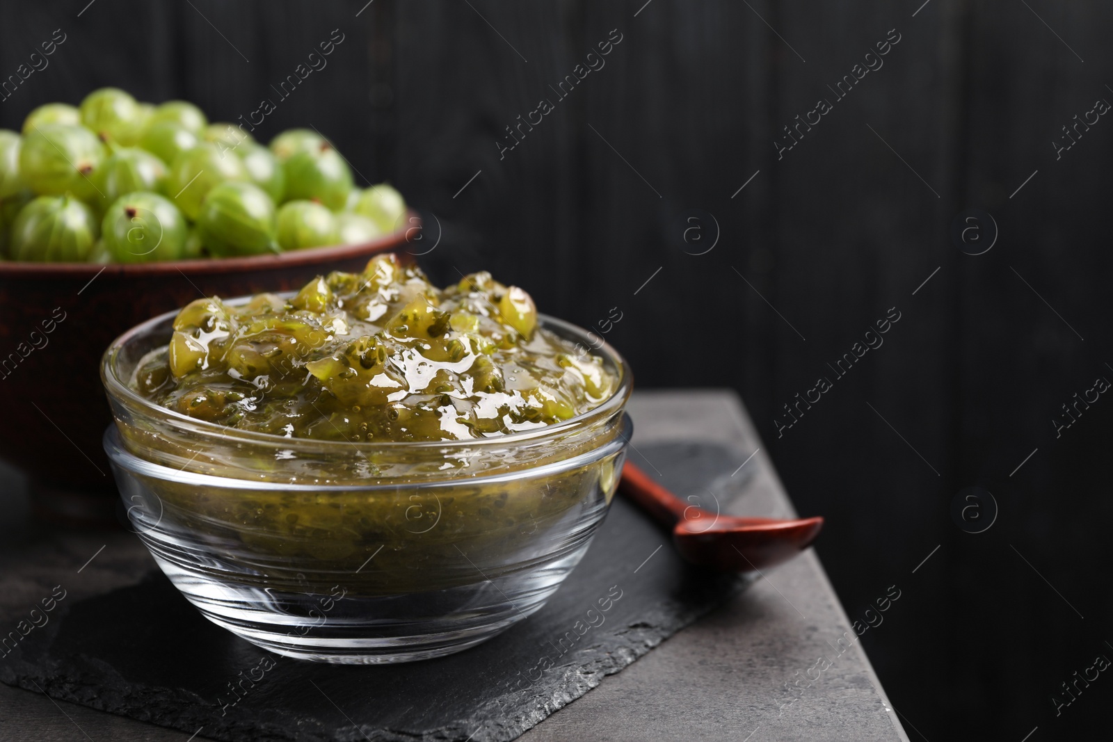 Photo of Delicious gooseberry jam in glass bowl and fresh berries on grey table, space for text