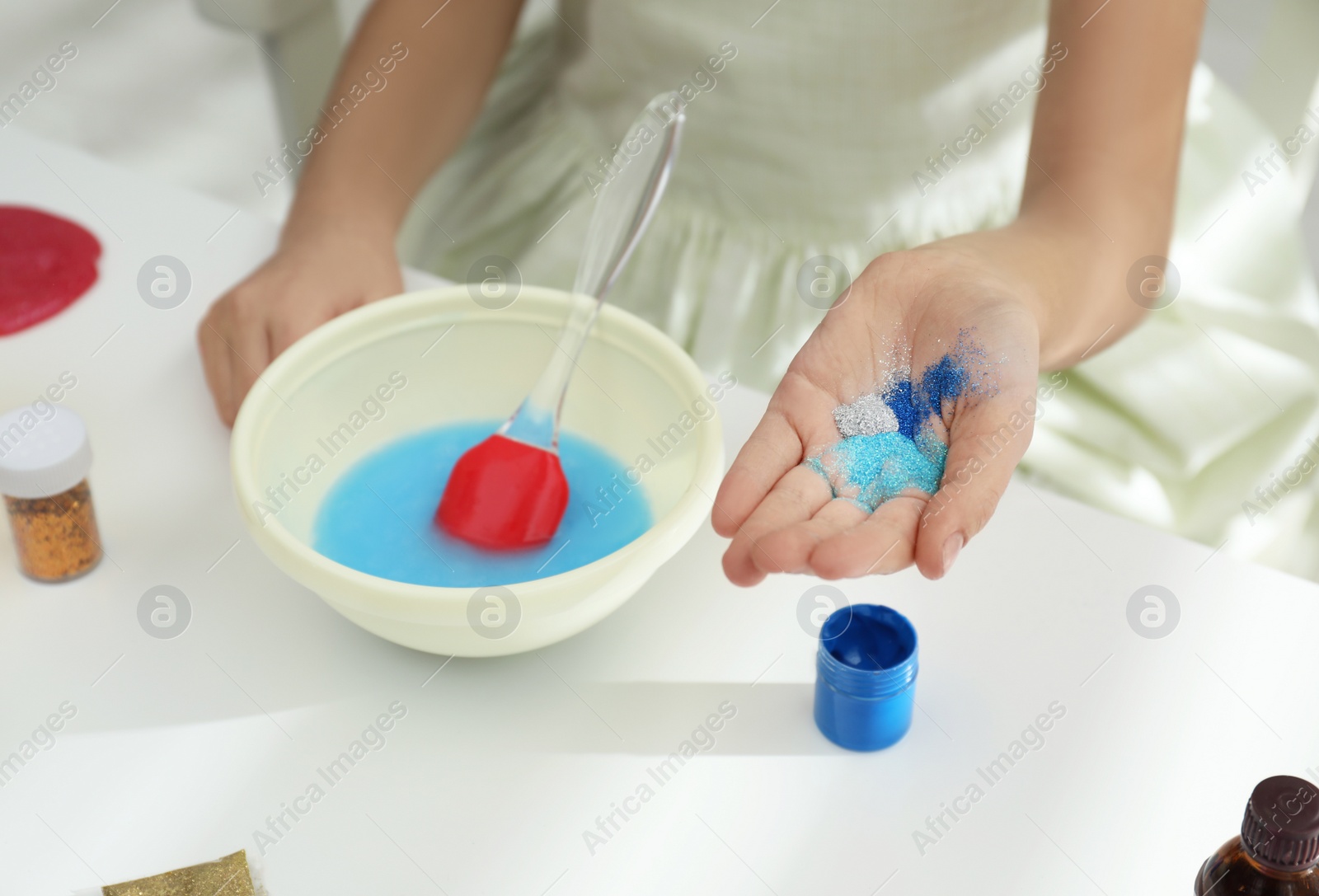 Photo of Little girl adding colored sparkles to homemade slime toy at table, closeup