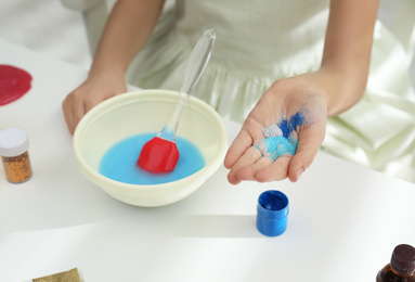 Little girl adding colored sparkles to homemade slime toy at table, closeup