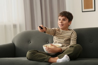 Photo of Little boy holding bowlpopcorn and changing TV channels with remote control on sofa at home