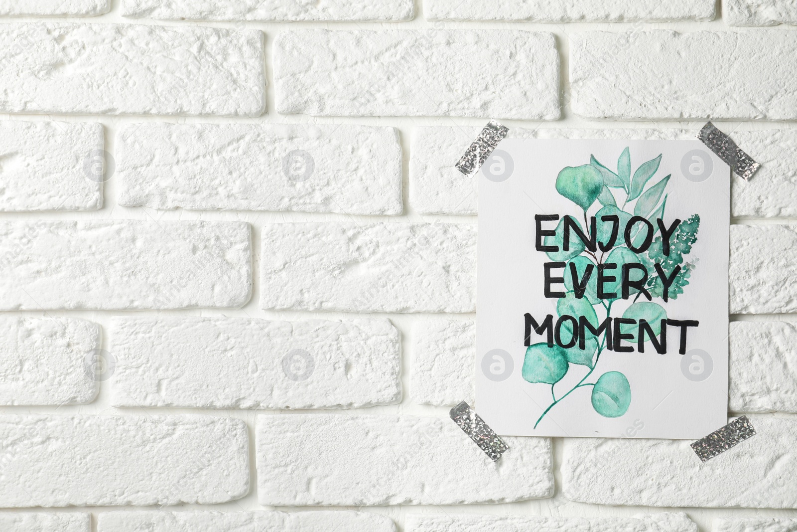 Photo of Card with phrase Enjoy Every Moment attached on white brick wall, space for text
