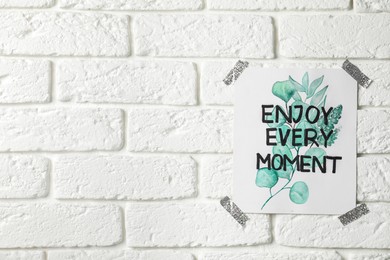 Photo of Card with phrase Enjoy Every Moment attached on white brick wall, space for text