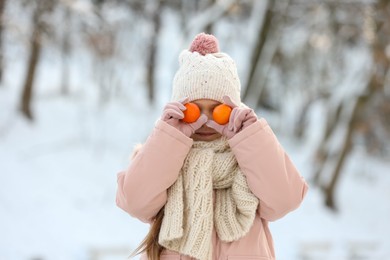 Cute little girl covering eyes with tangerines in snowy park on winter day