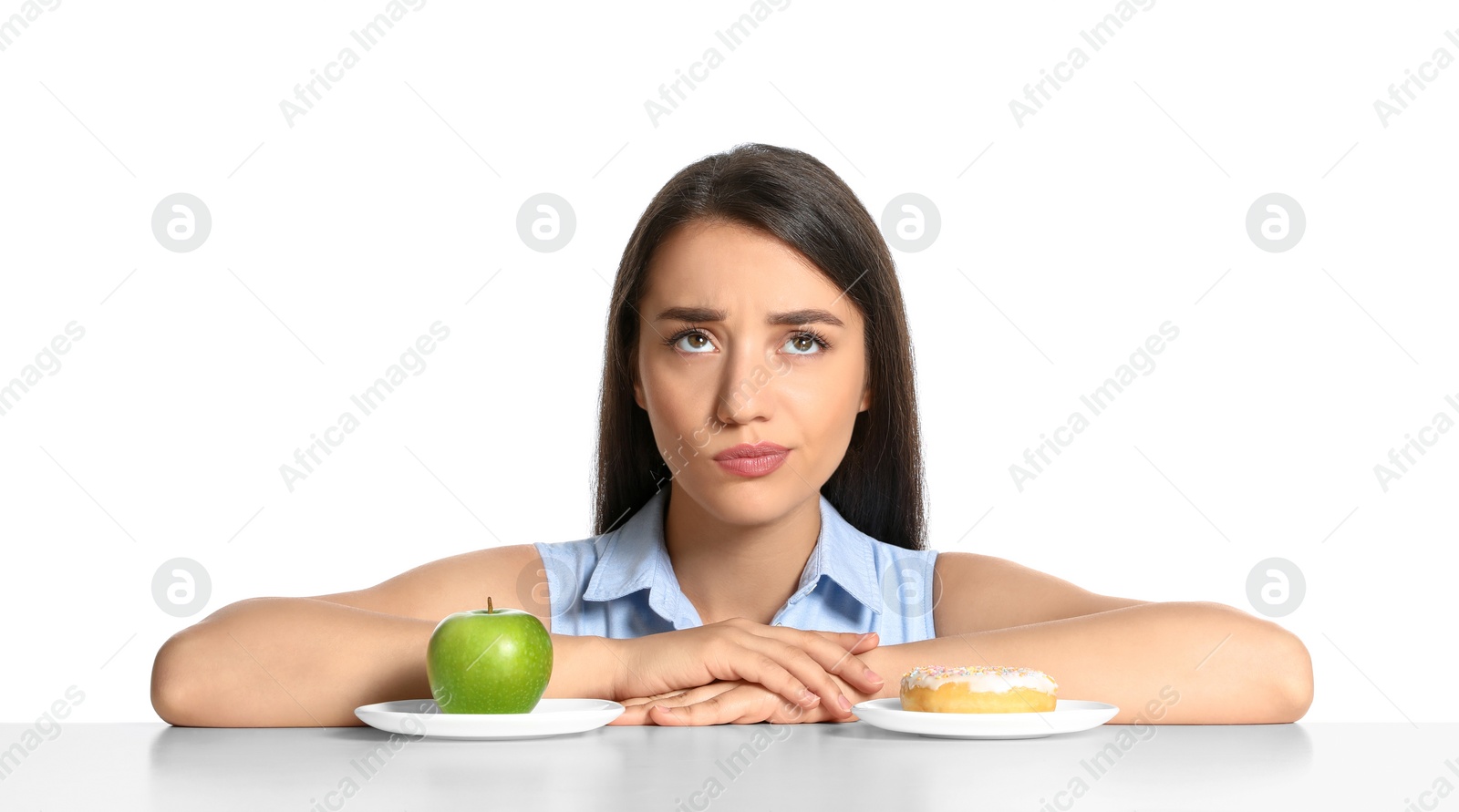 Photo of Doubtful woman choosing between apple and doughnut at table on white background