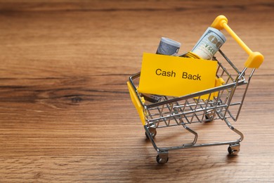 Photo of Card with word Cashback and rolled dollar banknotes in shopping cart on wooden background. Space for text