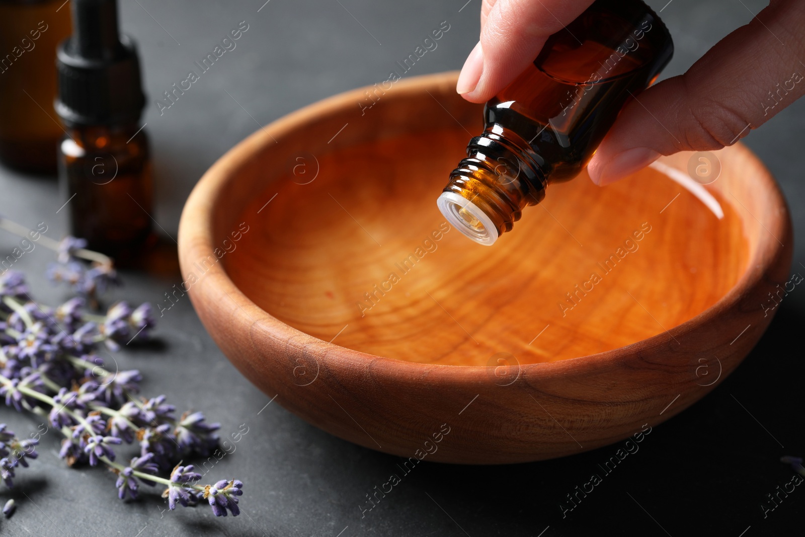 Photo of Woman dripping essential oil from bottle into bowl near lavender at grey table, closeup