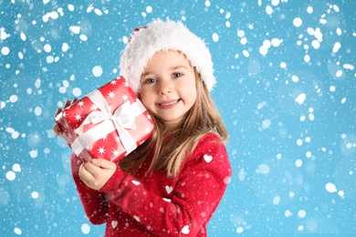 Image of Cute child in Santa hat with Christmas gift under snowfall on light blue background