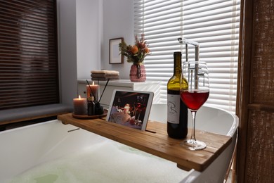 Photo of Wooden tray with tablet, wine and burning candles on bathtub in bathroom