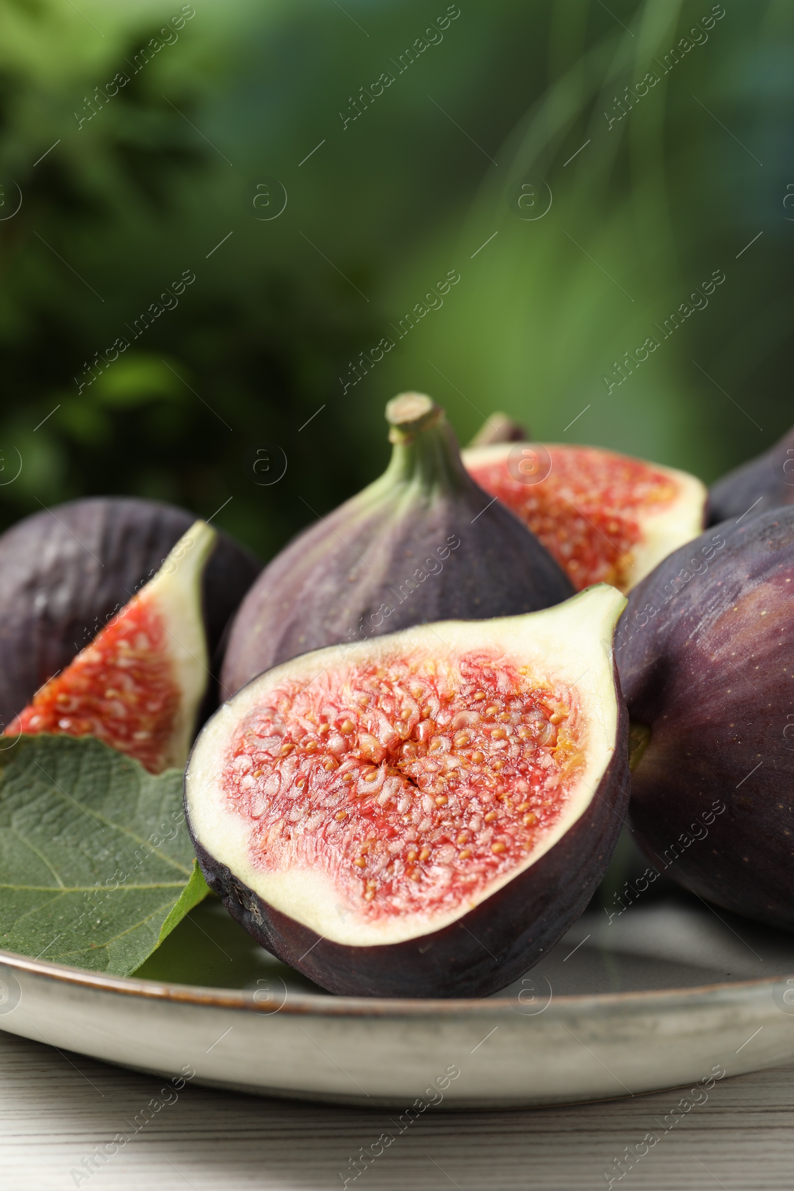 Photo of Whole and cut ripe figs with leaf on white wooden table against blurred green background, closeup