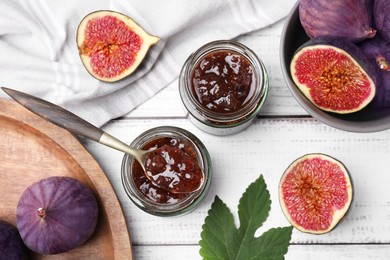 Glass jars of tasty sweet fig jam and fruits on white wooden table, flat lay