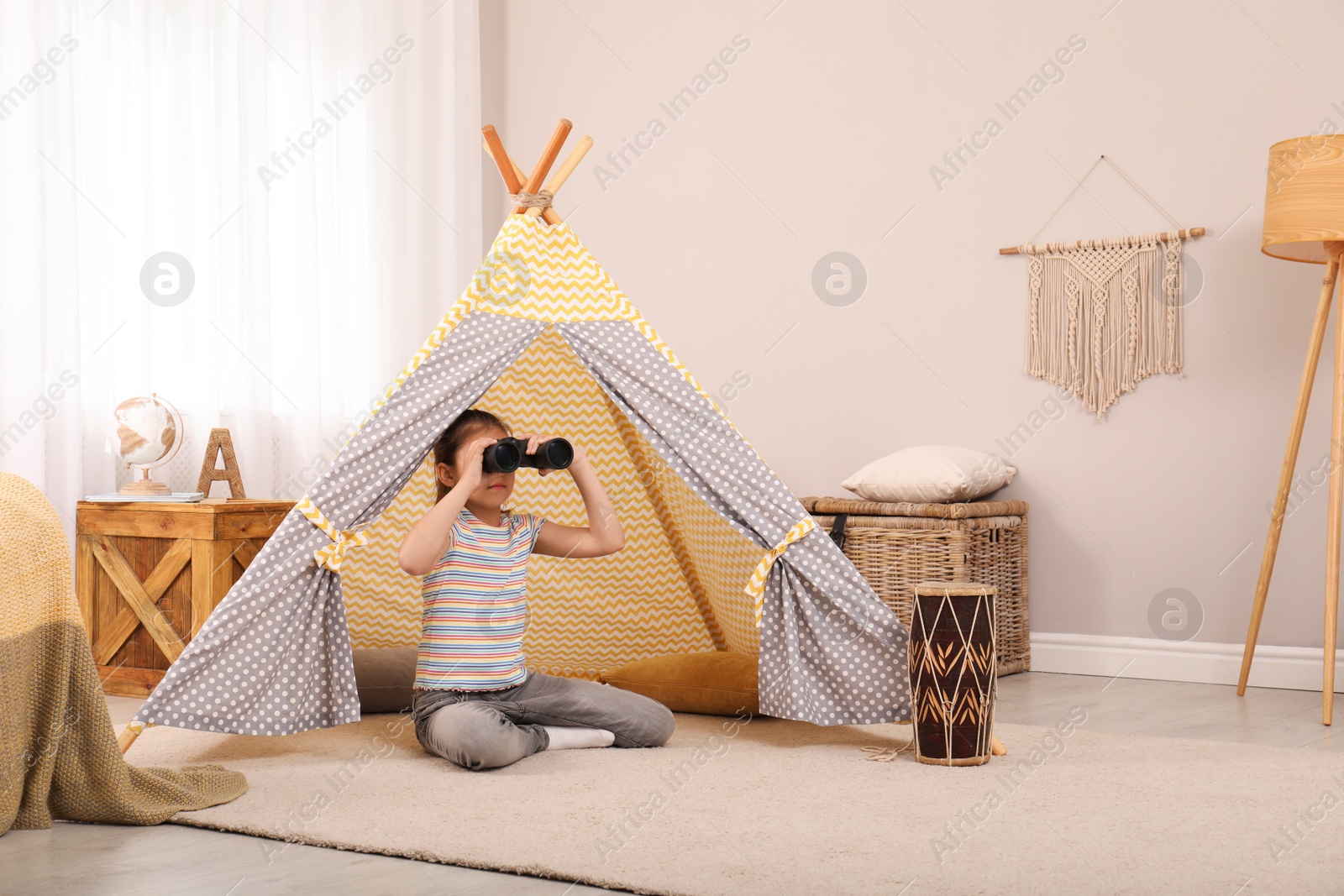 Photo of Cute little girl with binoculars in toy wigwam at home