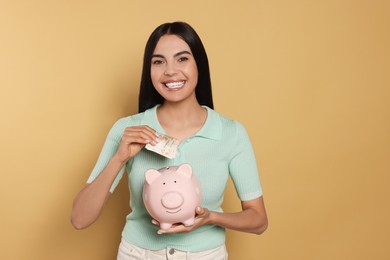 Photo of Happy young woman putting money into piggy bank on beige background