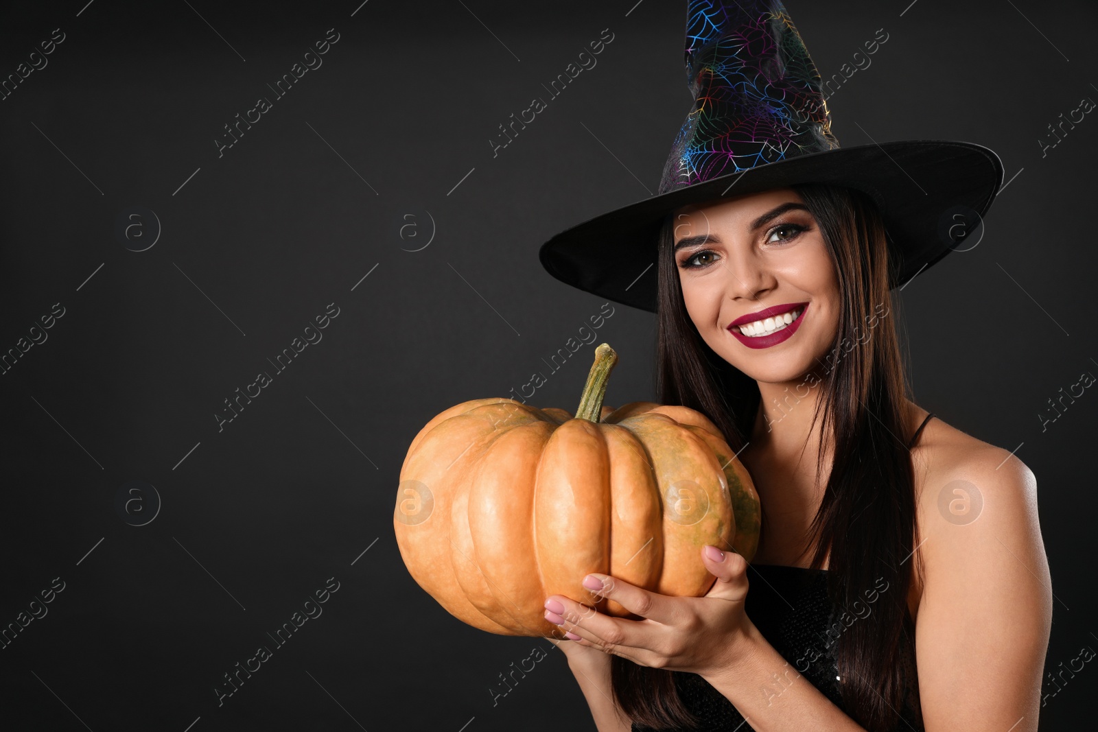 Photo of Beautiful woman wearing witch costume with pumpkin for Halloween party on black background, space for text