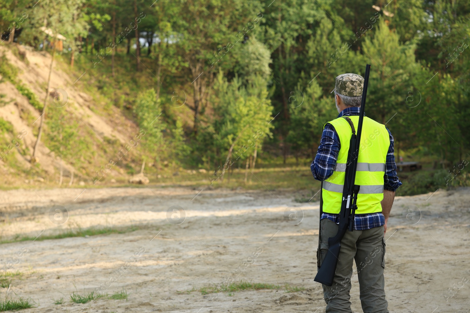 Photo of Man with hunting rifle wearing safety vest outdoors, back view. Space for text