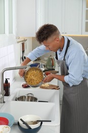 Photo of Man draining water from saucepan with pasta in messy kitchen. Many dirty dishware and utensils on countertop