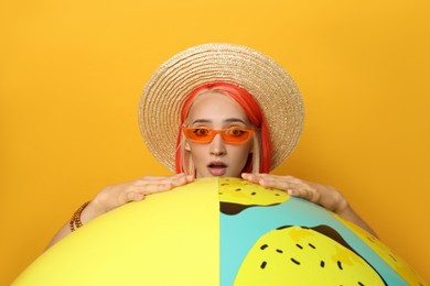 Emotional young woman with bright dyed hair and inflatable ball on orange background