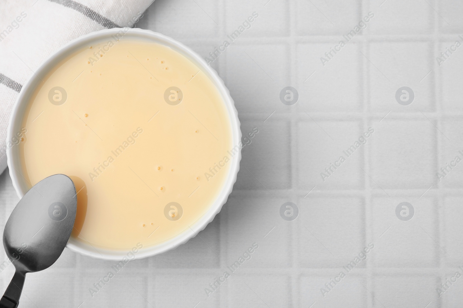 Photo of Bowl with condensed milk and spoon on white tiled table, flat lay. Space for text